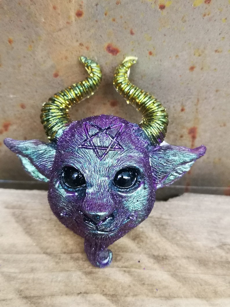 Puede incluir: Una figurita de resina iridiscente de color morado y verde de un peque&ntilde;o demonio con cuernos dorados y un pentagrama en la frente.