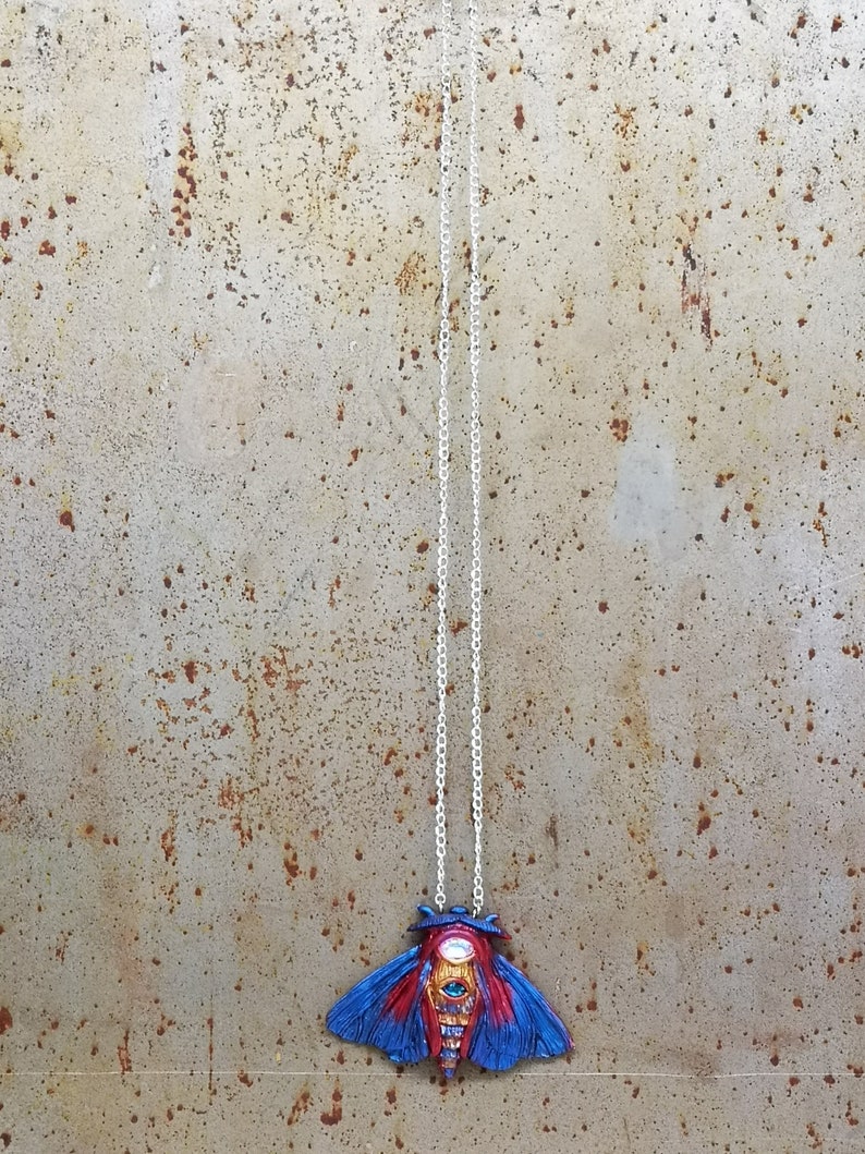 May include: A silver chain necklace with a blue and red moth pendant. The moth has a large, detailed eye with gold accents.