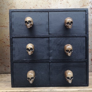May include: Black wooden chest of drawers with six drawers. Each drawer has a skull-shaped knob.