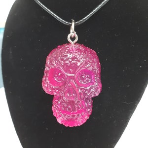 May include: A pink resin skull pendant necklace with intricate detailing. The pendant is attached to a black cord necklace.