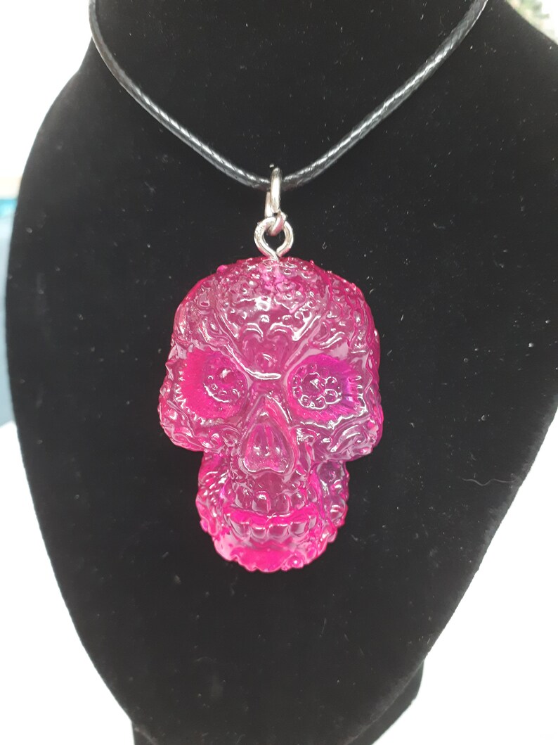 May include: A pink resin skull pendant necklace with intricate detailing. The pendant is attached to a black cord necklace.