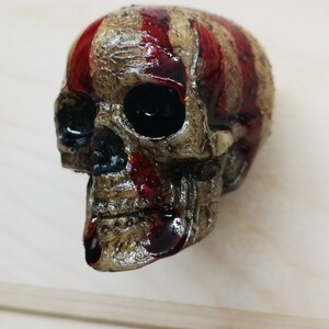 May include: A white skull with red stripes painted on the top of the head. The skull is made of resin and has a distressed finish.
