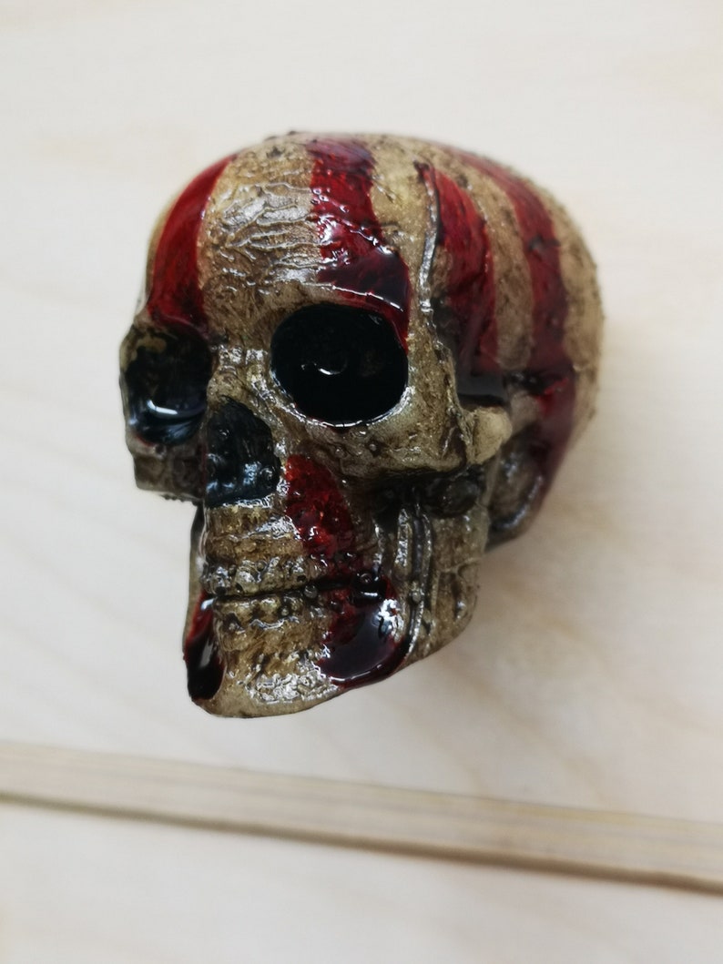 May include: A white skull with red stripes painted on the top of the head. The skull is made of resin and has a distressed finish.