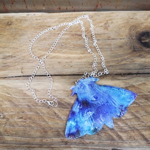 May include: A silver chain necklace with a blue and purple resin moth pendant. The moth has a detailed, textured design and iridescent wings.