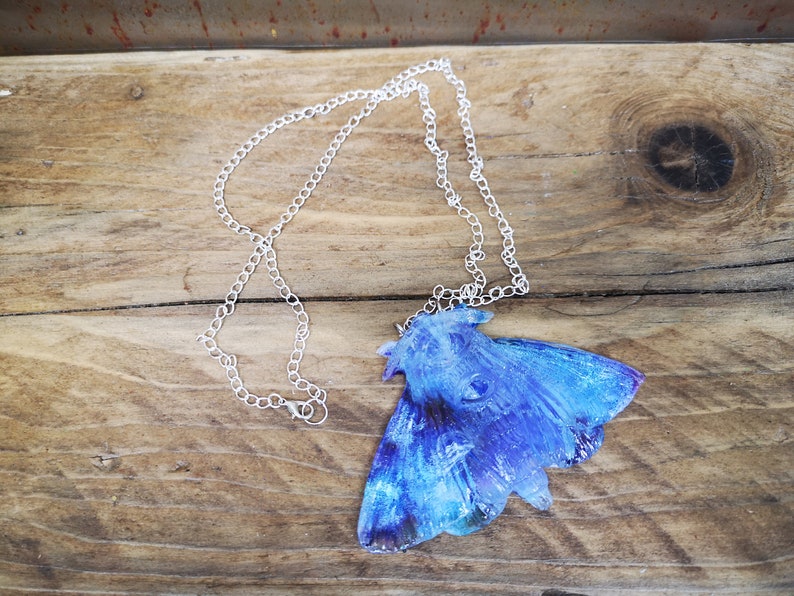 May include: A silver chain necklace with a blue and purple resin moth pendant. The moth has a detailed, textured design and iridescent wings.