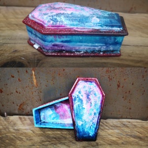 May include: A blue and pink coffin-shaped trinket box with a red glitter trim. The box is open and the inside is visible. The box is made of resin and has a unique, marbled appearance.