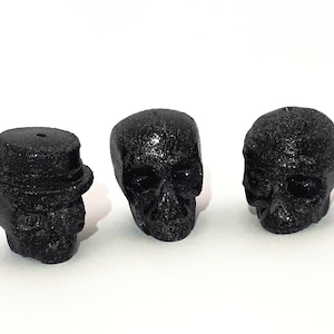 May include: Three black skull-shaped candle holders. The candle holders are made of a textured material and have a matte finish.