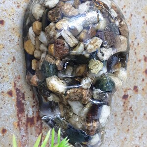 May include: A clear resin skull filled with small stones in shades of brown, white, and green.
