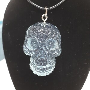 May include: A clear glass skull pendant necklace with intricate detailing. The skull is a traditional Day of the Dead design with a black cord.