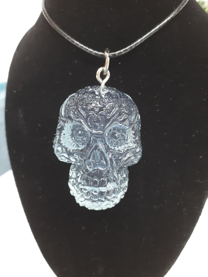 May include: A clear glass skull pendant necklace with intricate detailing. The skull is a traditional Day of the Dead design with a black cord.