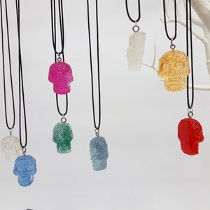 May include: Seven colorful sugar skull pendants on black cords. The skulls are made of clear resin and have a variety of colors, including pink, green, blue, red, yellow, and white.