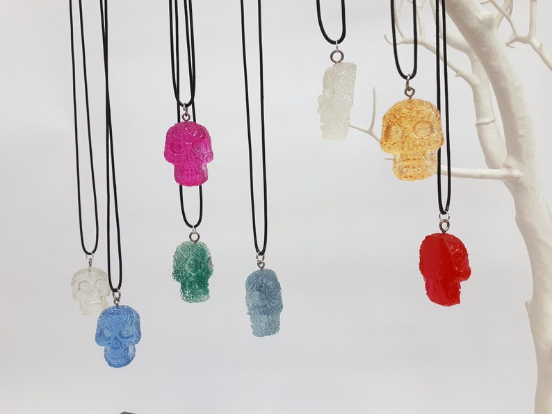 May include: Seven colorful sugar skull pendants on black cords. The skulls are made of clear resin and have a variety of colors, including pink, green, blue, red, yellow, and white.