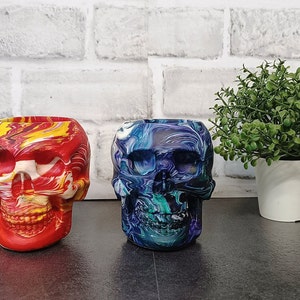 May include: Two colorful skull-shaped planters, one red and yellow with white swirls, the other blue and green with white swirls, sitting on a black surface in front of a white brick wall and a potted green plant.