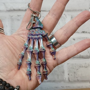 May include: A colourful skeleton hand keyring with a silver metal ring. The hand is painted in shades of purple, green, and brown.