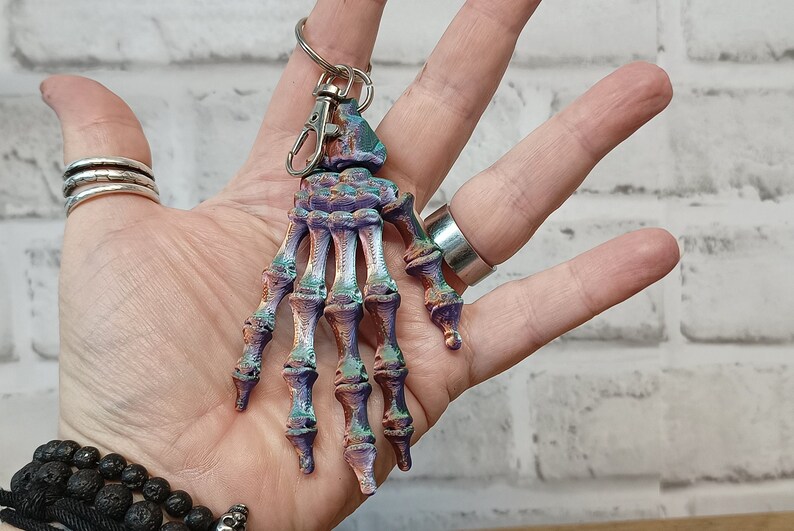 May include: A colourful skeleton hand keyring with a silver metal ring. The hand is painted in shades of purple, green, and brown.