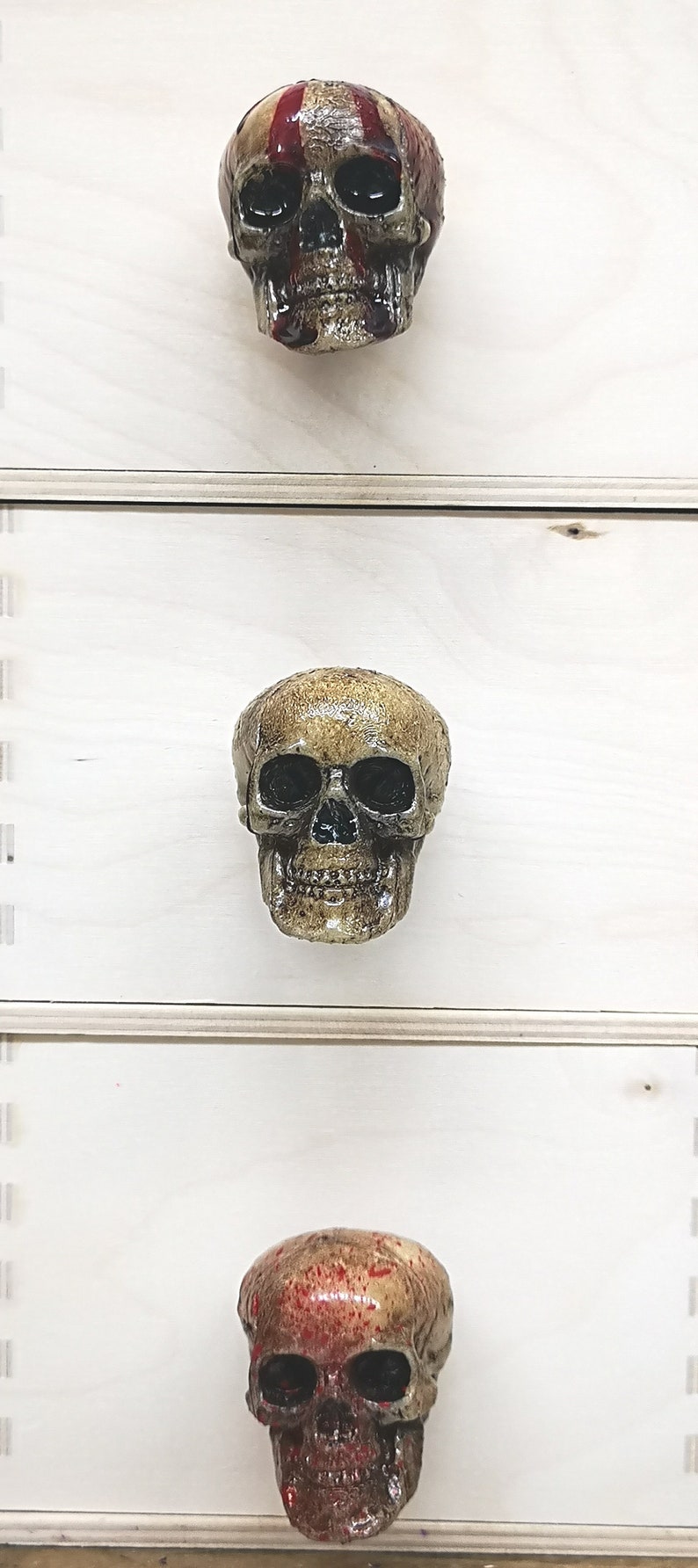 May include: Three skull-shaped drawer pulls, each with a different paint treatment. The top pull has red and white stripes painted on the skull. The middle pull is a plain, unpainted skull. The bottom pull has red splatter paint on the skull.