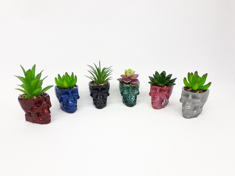 Pode incluir: Seis pequenos vasos com formato de caveira, cada um com uma cor diferente e uma pequena planta suculenta dentro. Os vasos s&atilde;o feitos de um material texturizado e t&ecirc;m um acabamento brilhante. As cores dos vasos s&atilde;o vermelho, azul, preto, verde, rosa e cinza.