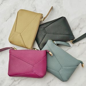 May include: Four small leather coin purses in different colors: mustard yellow, black, pink, and light blue. Each purse has a zipper closure and a geometric design.