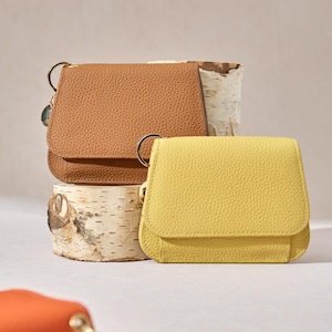 May include: A collection of small, textured leather wallets in shades of brown, yellow, and orange. Each wallet features a flap closure and a gold-tone ring for attaching a key or charm. The wallets are displayed on birch bark.