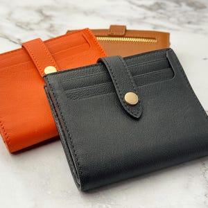 May include: Three leather wallets in different colors: orange, black, and brown. The wallets have a simple design with a flap closure and a gold button.