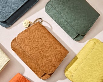 May include: A selection of colourful leather wallets in shades of blue, green, yellow, orange, and tan. Each wallet has a flap closure and a textured surface. One wallet has a gold-coloured keychain with a small charm.