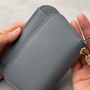 Op de afbeelding: Een grijze leren portemonnee met een gouden sleutelhanger eraan. De portemonnee heeft een gestructureerd oppervlak en is dubbelgevouwen.