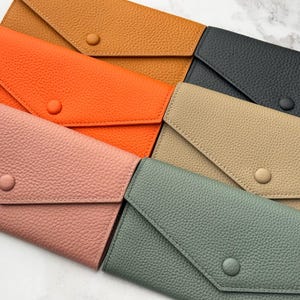 May include: A selection of leather wallets in shades of orange, pink, tan, and black. Each wallet has a flap closure with a button. The wallets are arranged diagonally, displaying their textured surfaces. The wallets are made of leather.