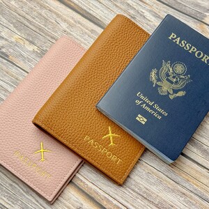 May include: Three passport covers, one brown, one pink, and one blue. The blue cover is a US passport with the words "PASSPORT" and "United States of America" printed on it. The brown and pink covers have the word "PASSPORT" printed on them.