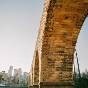 May include: A stone arch bridge with weathered, tan-colored masonry blocks spans across a frozen river. In the background, a city skyline with multiple high-rise buildings is visible under a clear, bright sky during the daytime.