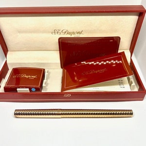 Puede incluir: Un bol&iacute;grafo dorado con superficie texturizada en una caja roja con el logotipo de S.T. Dupont y una tarjeta de garant&iacute;a. La caja est&aacute; abierta y el bol&iacute;grafo est&aacute; encima de la caja.