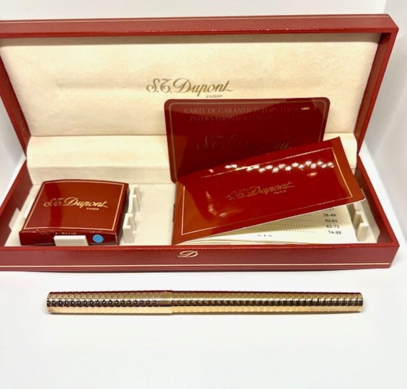 Puede incluir: Un bol&iacute;grafo dorado con superficie texturizada en una caja roja con el logotipo de S.T. Dupont y una tarjeta de garant&iacute;a. La caja est&aacute; abierta y el bol&iacute;grafo est&aacute; encima de la caja.