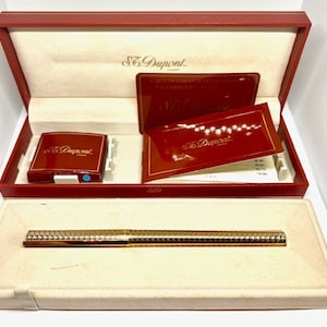 Puede incluir: Un bol&iacute;grafo S.T. Dupont de color dorado en su caja roja con una caja roja m&aacute;s peque&ntilde;a y papeles. El bol&iacute;grafo tiene un dise&ntilde;o texturizado.