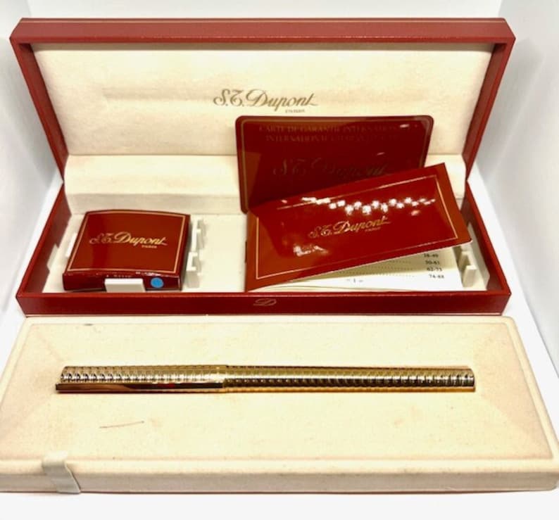 Puede incluir: Un bol&iacute;grafo S.T. Dupont de color dorado en su caja roja con una caja roja m&aacute;s peque&ntilde;a y papeles. El bol&iacute;grafo tiene un dise&ntilde;o texturizado.