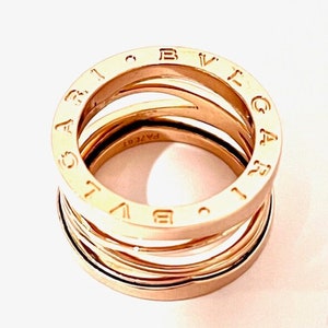 May include: A gold-toned ring with the word "BVLGARI" engraved around the band. The ring is made of multiple bands that are stacked on top of each other.