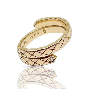 May include: A gold-colored ring with a quilted design and two small diamonds. The ring is marked with "INEL" and "Au 750". The ring is shown on a white background with a reflection.
