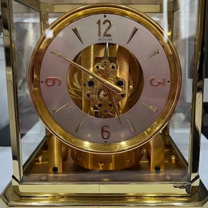 May include: A gold-toned, square-shaped clock with a glass face. The clock face has a visible, intricate mechanism with gold-toned hands and Roman numeral markings. The clock face also has the text "LeCoultre" printed on it.