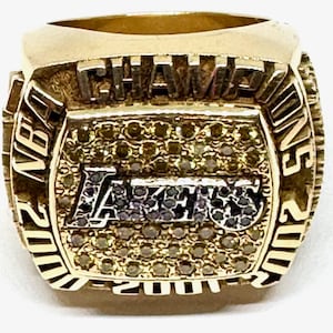 May include: A gold NBA championship ring with the words "NBA Champions 2000-2001" and "Los Angeles Lakers" on it. The ring is adorned with diamonds.