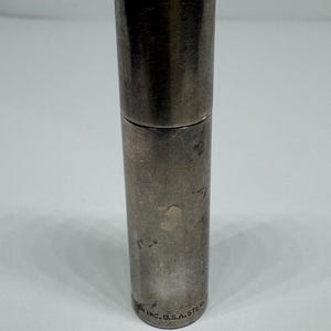 May include: A cylindrical, silver-toned metal object with a cap. The object is divided into two sections, with a slightly darker tone on the lower portion. The base of the object has the text "INC. U.S.A. STERL".