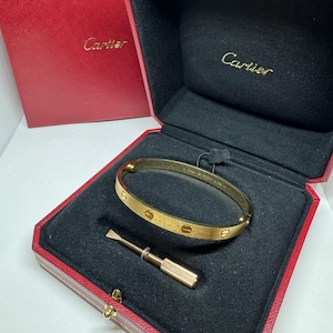 May include: A gold-colored bracelet with screw details and a matching screwdriver, presented in a red box with the brand name "Cartier" embossed on the lid and inside. The bracelet is on a black velvet interior.