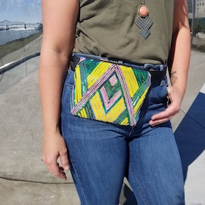 May include: A green, yellow and pink beaded bum bag with a geometric pattern. The bum bag is worn around the waist of a person wearing blue jeans.