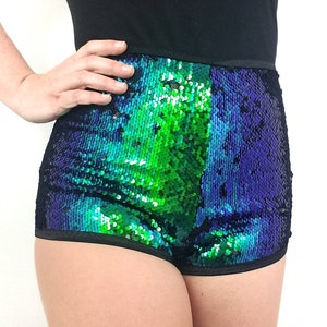 May include: High-waisted shorts covered in iridescent sequins, transitioning from green to blue. The shorts have a black waistband and trim, creating a striking contrast. The garment is shown on a person with their hand on their hip.