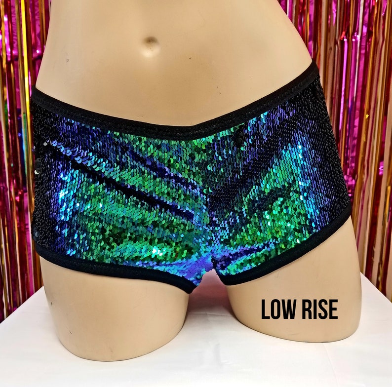 May include: Low-rise shorts with iridescent sequins in shades of green and blue, bordered with black trim. The shorts are displayed on a mannequin. The text "LOW RISE" is printed in black on the mannequin.