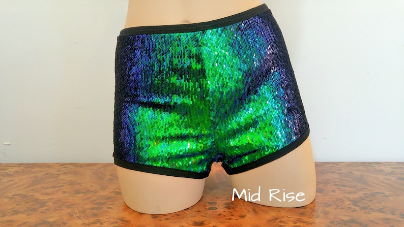 May include: Sequin shorts in a vibrant green and blue color scheme. The shorts have a black trim around the waist and leg openings. The mid-rise design is visible. The sequins create a shimmering effect.