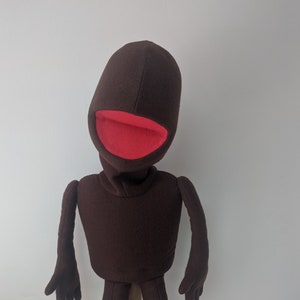 Könnte beinhalten: Eine braune Handpuppe aus Filz mit rotem Mund. Die Puppe steht auf einem Holzständer.