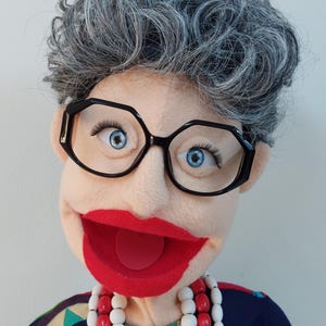 May include: A puppet with a light skin tone, grey curly hair, and large black-framed glasses. The puppet has blue eyes, long eyelashes, and a wide red mouth. It wears a necklace of red and white beads and a patterned top.