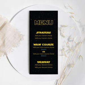 May include: A black menu card with yellow text, featuring the word "MENU" at the top. The menu is divided into sections for starters, main course, and dessert. The card is on a white plate, with a beige cloth and dried flowers in the background.