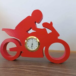 Könnte beinhalten: Eine rote Holz-Uhr in Form eines Motorrads mit Fahrer. Das Zifferblatt ist rund mit einem goldfarbenen Rahmen und schwarzen Ziffern. Die Zeiger zeigen auf etwa 6:00 Uhr.