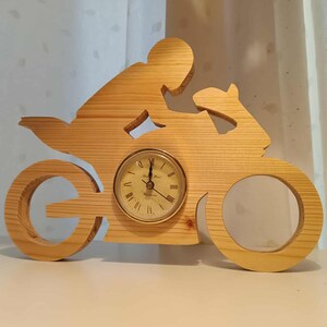 Könnte beinhalten: Holzuhr in Motorradform mit rundem Zifferblatt. Die Uhr zeigt eine Fahrer-Silhouette und Radeinschnitte. Das Zifferblatt hat schwarze römische Ziffern und goldene Zeiger. Die Uhr ist aus hellem Holz gefertigt.