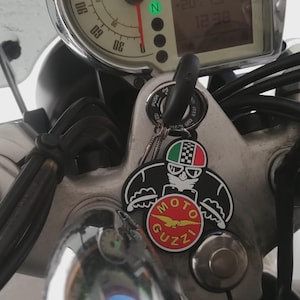 May include: Close-up of a motorcycle's dashboard and handlebars. A key with a Moto Guzzi keychain hangs from the ignition. The keychain features a cartoon character wearing a helmet with the Italian flag colors and the Moto Guzzi logo.