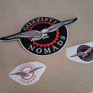 Puede incluir: Tres parches bordados con un diseño de águila en blanco y negro. Los parches muestran el texto "Guzzisti Nomads" en blanco sobre fondo negro. El parche más grande tiene un diseño de engranaje rojo.